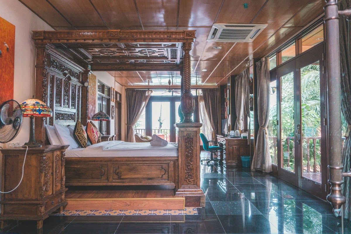 A suite with an ornate carved-teak four-poster bed, Tiffany lamp, timber ceiling and balcony doors opening to tropical greenery